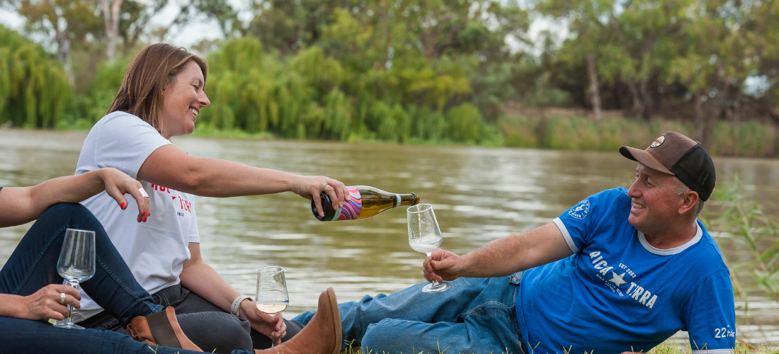 Man and woman smiling and pouring wine while laying in front of a river